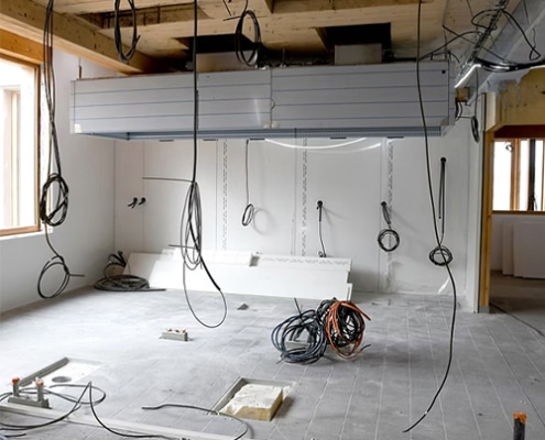 Unfinished interior under construction with exposed electrical cables hanging from the ceiling and building materials across the floor.