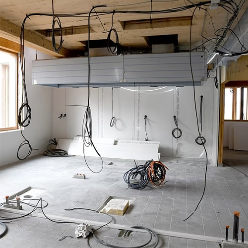 Unfinished interior under construction with exposed electrical cables hanging from the ceiling and building materials across the floor.