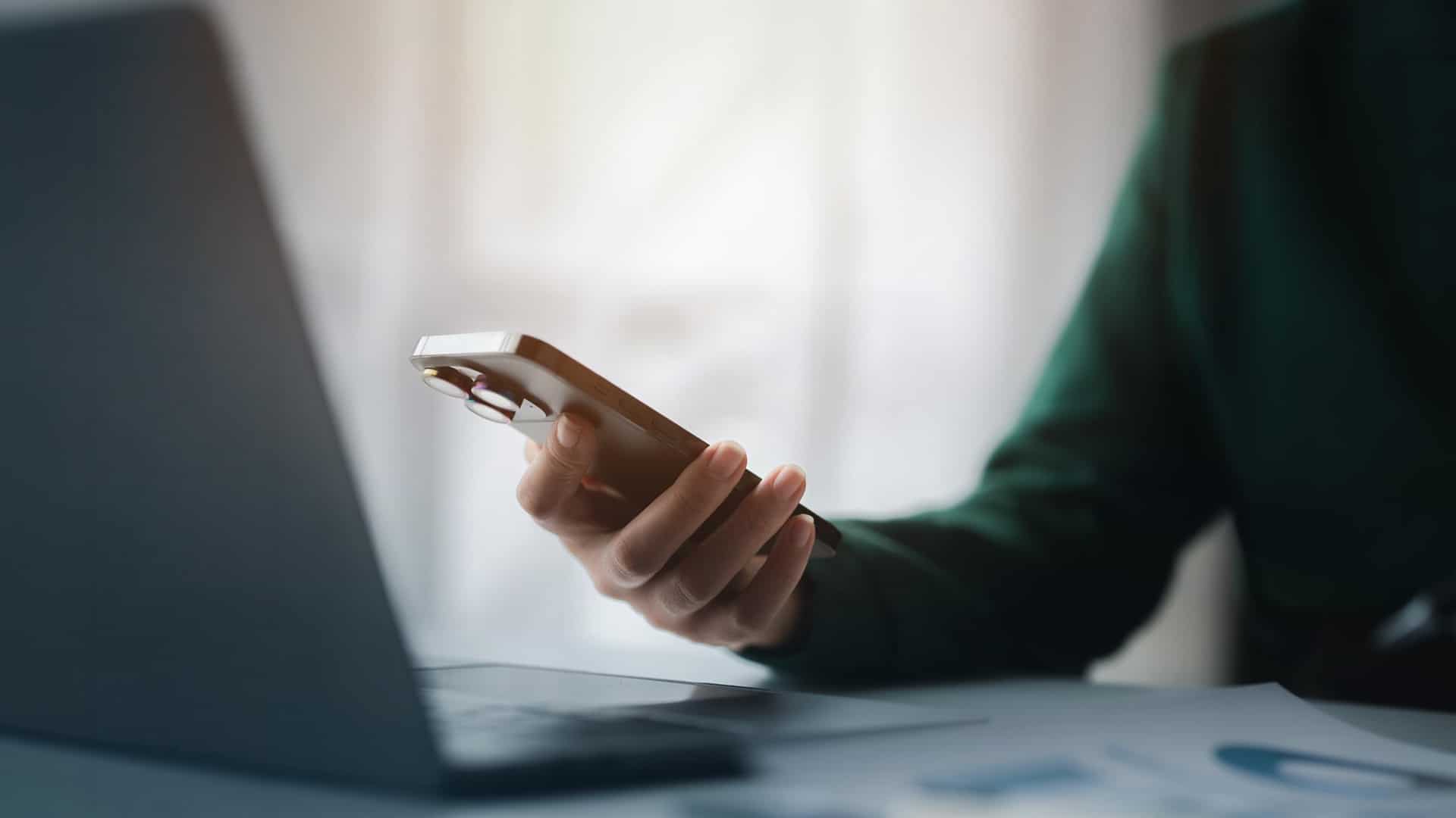 About > Reviews > Header > Background Person looking at online reviews on a smartphone beside a laptop.