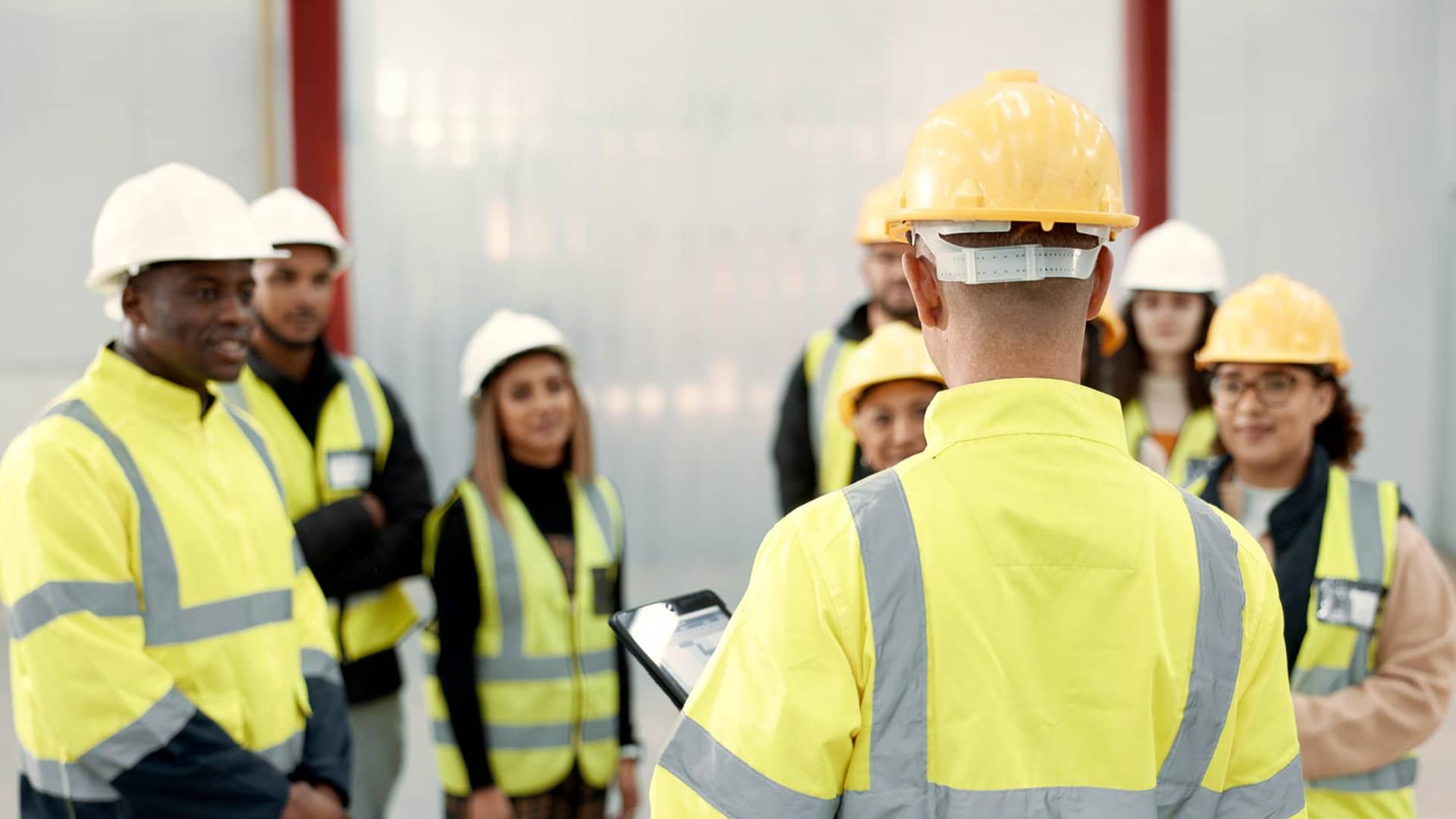 About > Who We Are > Header > Background Trainer delivering a health and safety briefing to construction workers wearing hard hats and high-visibility clothing.
