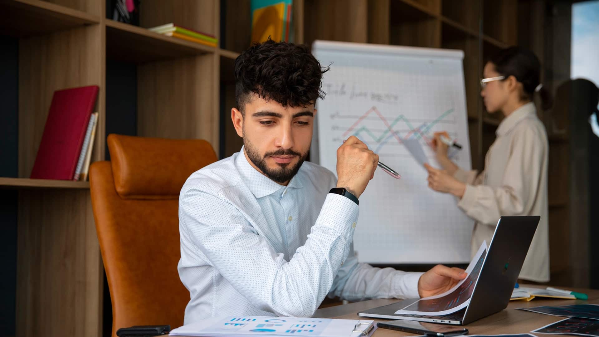 Courses > Business Skills > Header > Background Business professional reviewing data on a laptop while a colleague presents charts in the background.