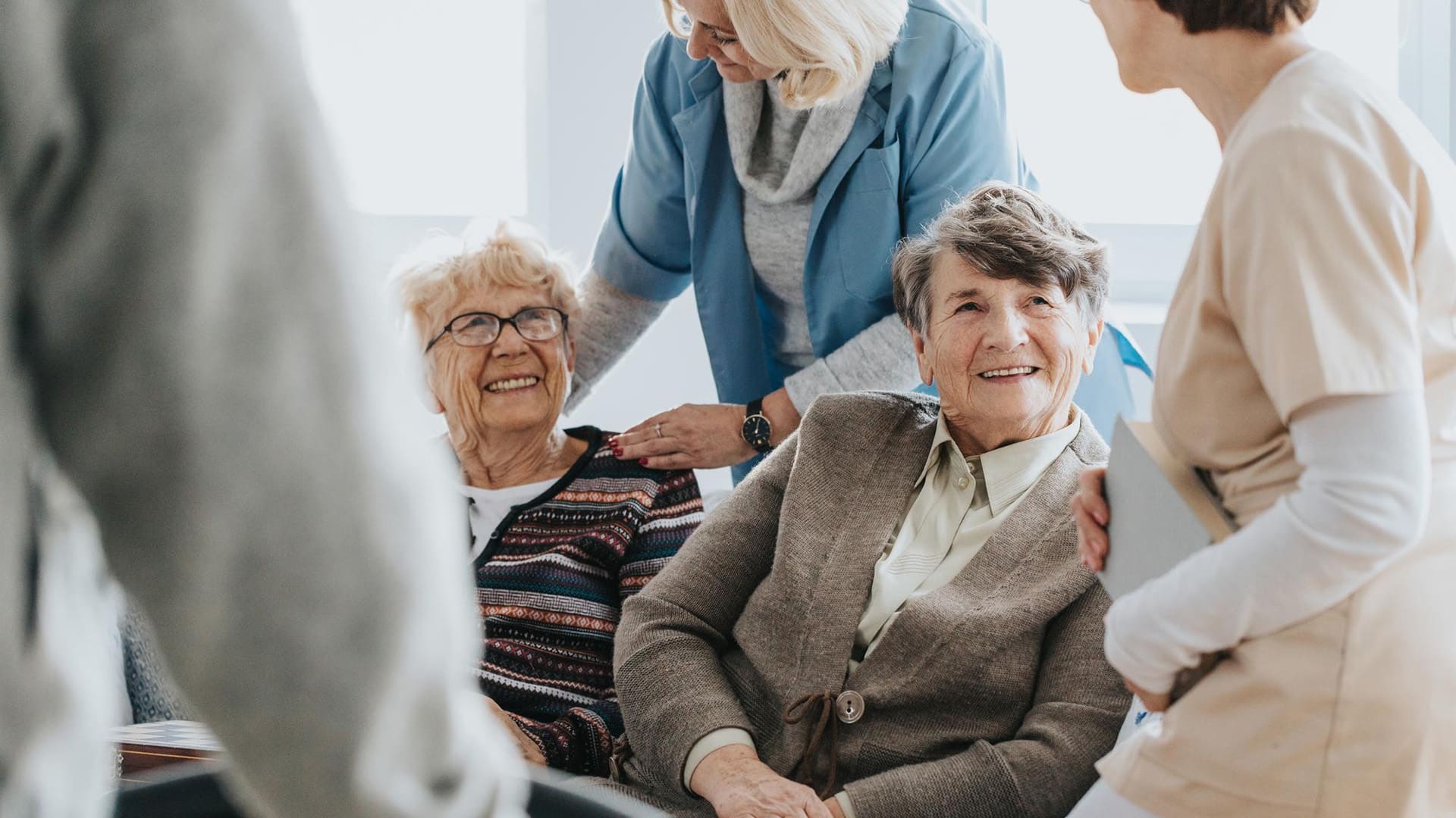 Courses > Care Homes > Header > Background Care workers supporting and speaking with elderly residents in a bright communal setting.