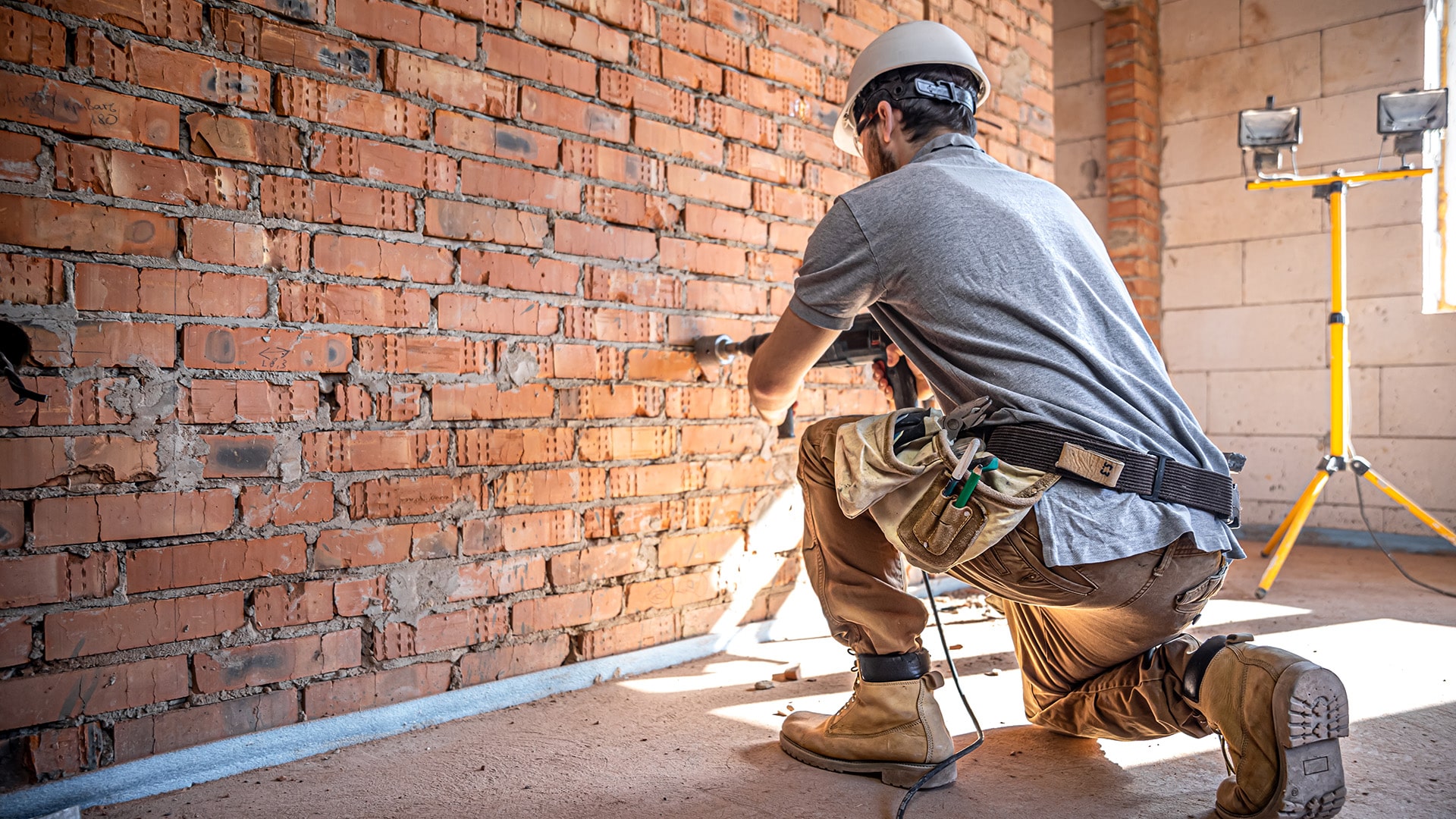 Courses > Construction > Header > Background Construction worker wearing a hard hat and tool belt while working on an exposed brick wall.