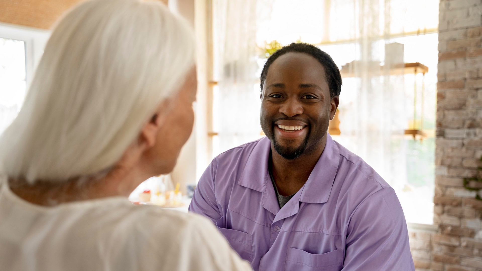 Courses > Health and Social Care > Header > Background Care worker smiling while speaking with an elderly woman in a bright residential setting.