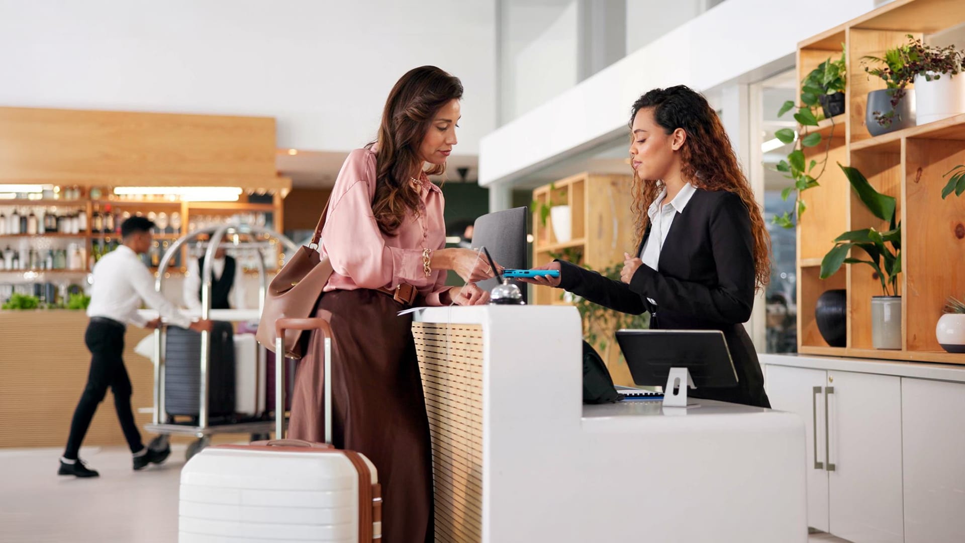 Courses > Hospitality > Header > Background Hotel receptionist assisting a guest checking in with luggage at the front desk.