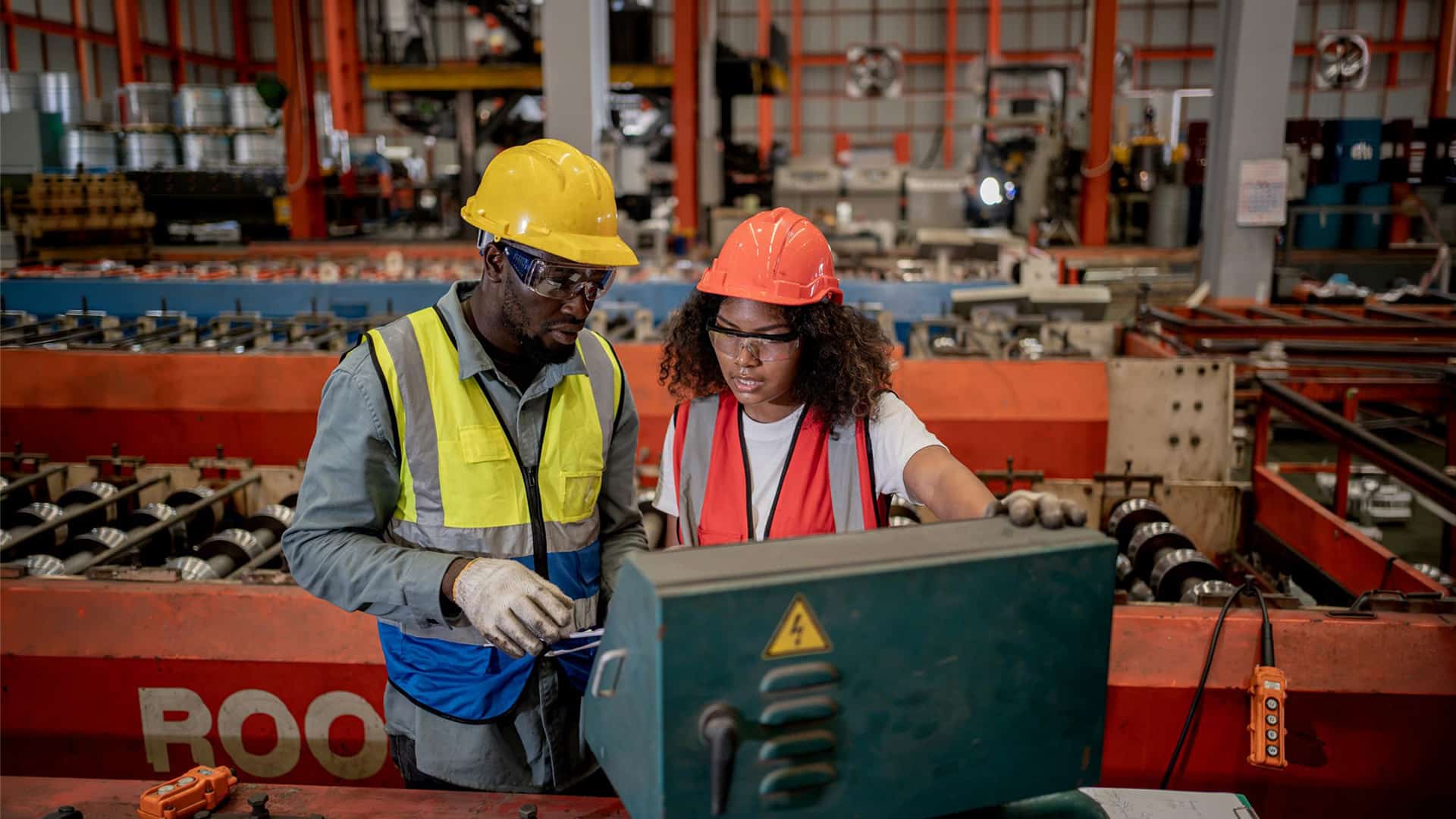 NVQs > Header > Background Two factory workers wearing hard hats, safety glasses and high-visibility vests operating industrial machinery.
